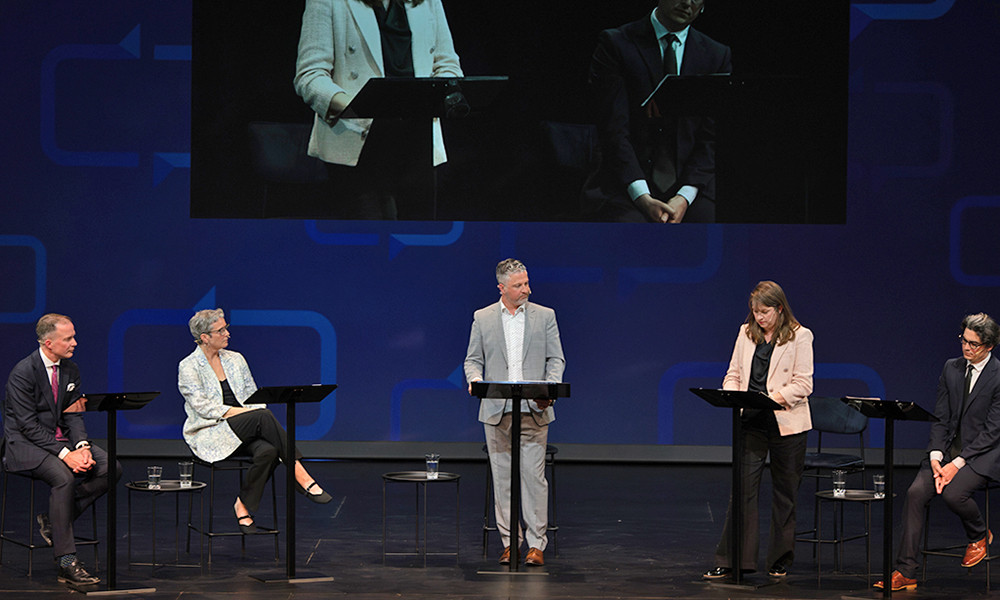 Five panelists seated and standing on stage during a live debate event. A projection screen above shows a close-up of the speakers.